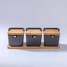 Load image into Gallery viewer, Ceramic Seasoning Storage Jars by Allthingscurated are the perfect storage solution to keep your herbs, seasonings and spices. Made of durable ceramic with a sustainable bamboo flip lid for easy access during cooking. These jars are not only functional but also environmentally friendly. Comes with a bamboo tray and ceramic spoons. Featured here is a set of 3 black jars with spoons and tray.