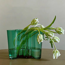 Load image into Gallery viewer, Green glass vase with green floral on a wooden surface. The Dayton Vase by Allthingscurated offers a beautiful asymmetrical design that feels both timeless and organic. Its sculptural, minimalist form brings a striking yet harmonious touch to any home interior. Available in clear, green and amber, it’s an unforgettable gift for housewarmings, weddings or holidays.
