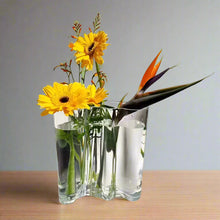 Load image into Gallery viewer, Clear glass vase with yellow flowers and a bird of paradise against a light gray background. The Dayton Vase by Allthingscurated offers a beautiful asymmetrical design that feels both timeless and organic. Its sculptural, minimalist form brings a striking yet harmonious touch to any home interior. Available in clear, green and amber, it’s an unforgettable gift for housewarmings, weddings or holidays.