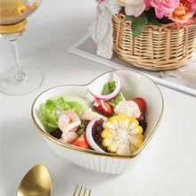 Load image into Gallery viewer, White heart-shaped bowl with salad on a table with a glass of wine and flowers. Gold-rimmed Heart Shape Ceramic Bowl by Allthingscurated is a beautiful and versatile piece to own in your dinnerware collection. Perfect for salads, desserts and appetizers, its charming design adds an extra touch of romance for Valentine’s Day dinner; and make everyday meals extra special.