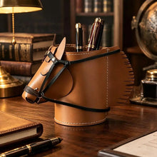Load image into Gallery viewer, A brown faux leather horse-shaped pen holder with black straps, shown on a tabletop with pens inserted inside. The scene includes styled props like vintage table lamp, hard cover books, and a notebook on a wooden table, in a softly lit indoor setting.