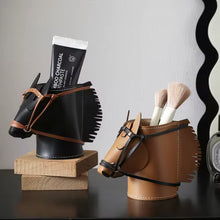 Load image into Gallery viewer, Two horse-shaped holders on a dark tabletop against a light gray wall. The left holder is black faux leather with brown straps and holds a black tube of toothpaste on a wooden base. The right holder is brown faux leather with brown straps and holds two cosmetic brushes.