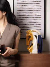 Load image into Gallery viewer, Person holding a cup next to a decorative horse head sculpture on a wooden surface. Jyunma Ceramic Horse by Allthingscurated is handmade artistry at its finest. It’s more than just decoration, it embodies auspiciousness and prosperity; represents steadfast loyalty and protective guardianship through time. Its elegance makes it a perfect gift for any horse enthusiasts or anyone born under the Chinese zodiac sign of the horse; and an exquisite addition to any contemporary home for its enduring appeal.