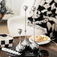 Load image into Gallery viewer, Set of three tall, silver candle holders on a wooden table with a blurred background. Lumea Silver Candle Holders by Allthingscurated feature a striking trio of sculptural pieces that blend contemporary design with timeless elegance. Each holder features a distinctive water droplet base that anchors a sleek, minimalist silhouette, creating an art deco-inspired statement on its own. this set makes an exceptional wedding gift for couples seeking sophisticated home décor that transcends occasion.