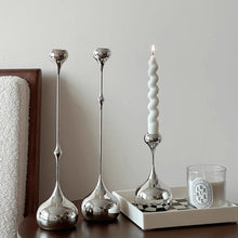 Load image into Gallery viewer, Three silver candle holders and a white candle on a wooden surface with a neutral background. Lumea Silver Candle Holders by Allthingscurated feature a striking trio of sculptural pieces that blend contemporary design with timeless elegance. Each holder features a distinctive water droplet base that anchors a sleek, minimalist silhouette, creating an art deco-inspired statement on its own. this set makes an exceptional wedding gift for couples seeking sophisticated home décor that transcends occasion.