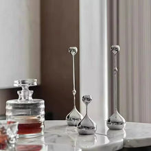 Load image into Gallery viewer, Set of silver candle holders on a marble surface with a neutral background. Lumea Silver Candle Holders by Allthingscurated feature a striking trio of sculptural pieces that blend contemporary design with timeless elegance. Each holder features a distinctive water droplet base that anchors a sleek, minimalist silhouette, creating an art deco-inspired statement on its own. this set makes an exceptional wedding gift for couples seeking sophisticated home décor that transcends occasion.
