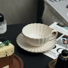 Load image into Gallery viewer, Off-white ceramic cup and saucer on a dark surface with a small cake and decorative items. Japanese Chrysanthemum Cup and Saucer by Allthingscurated is inspired by the flower’s perfect symmetry and stunning beauty in the Japanese culture. Crafted from porcelain, it’s a beautiful set to entertain friends and family for afternoon teas at home.