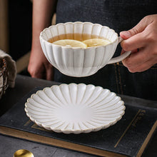 Load image into Gallery viewer, Person holding a white ceramic teacup with a scalloped edge, filled with tea, on a dark surface. Japanese Chrysanthemum Cup and Saucer by Allthingscurated is inspired by the flower’s perfect symmetry and stunning beauty in the Japanese culture. Crafted from porcelain, it’s a beautiful set to entertain friends and family for afternoon teas at home.