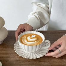 Load image into Gallery viewer, Person holding a off-white ceramic cup set with latte art on a wooden table. Japanese Chrysanthemum Cup and Saucer by Allthingscurated is inspired by the flower’s perfect symmetry and stunning beauty in the Japanese culture. Crafted from porcelain, it’s a beautiful set to entertain friends and family for afternoon teas at home.