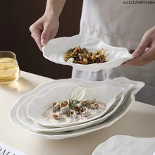 Load image into Gallery viewer, Person holding a white oyster-shaped dish with food on a table. Oyster Shape Serving Dish by Allthingscurated is elegantly crafted in pristine white ceramic to mirror the contours and texture of the elegant oyster. The striking serve ware comes in multiple sizes for plating appetizers or main courses.
