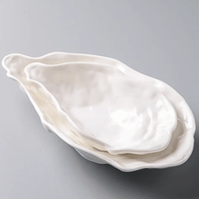 Load image into Gallery viewer, Set of two white oyster shape ceramic dishes on a gray background. Oyster Shape Serving Dish by Allthingscurated is elegantly crafted in pristine white ceramic to mirror the contours and texture of the elegant oyster. The striking serve ware comes in multiple sizes for plating appetizers or main courses.