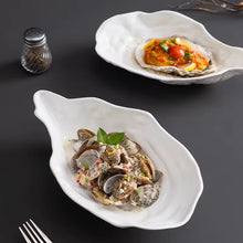 Load image into Gallery viewer, Two white oyster shapenceramic dishes with seafood on a dark surface. Oyster Shape Serving Dish by Allthingscurated is elegantly crafted in pristine white ceramic to mirror the contours and texture of the elegant oyster. The striking serve ware comes in multiple sizes for plating appetizers or main courses.