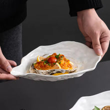 Load image into Gallery viewer, Person holding a white oyster dish with a cooked oyster garnished with lemon and herbs on a dark background. Oyster Shape Serving Dish by Allthingscurated is elegantly crafted in pristine white ceramic to mirror the contours and texture of the elegant oyster. The striking serve ware comes in multiple sizes for plating appetizers or main courses.