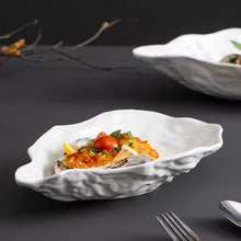Load image into Gallery viewer, Oyster shape dish with a cooked oyster garnished with vegetables on a dark surface. Oyster Shape Serving Dish by Allthingscurated is elegantly crafted in pristine white ceramic to mirror the contours and texture of the elegant oyster. The striking serve ware comes in multiple sizes for plating appetizers or main courses.