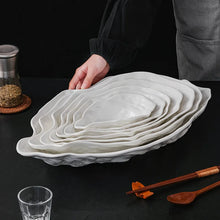 Load image into Gallery viewer, Stack of white oyster shape ceramic plates on a dark surface with a person's hand and kitchen items in the background. Oyster Shape Serving Dish by Allthingscurated is elegantly crafted in pristine white ceramic to mirror the contours and texture of the elegant oyster. The striking serve ware comes in multiple sizes for plating appetizers or main courses.