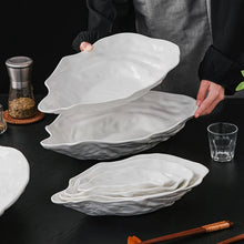 Load image into Gallery viewer, White ceramic oyster-shaped plates on a dark surface with a person holding one plate. Oyster Shape Serving Dish by Allthingscurated is elegantly crafted in pristine white ceramic to mirror the contours and texture of the elegant oyster. The striking serve ware comes in multiple sizes for plating appetizers or main courses.