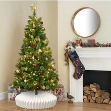Load image into Gallery viewer, Decorated Christmas tree with lights, ornaments and white paper collar in a living room with a fireplace and stockings. Transform your holiday setup with the striking Honeycomb Christmas Tree Collar by Allthingscurated. Crafted from premium paper with easy setup, its distinctive pleated design delivers sophisticated visual interest while concealing the tree base with understated elegance. The crips white finish works beautifully with any tree style.