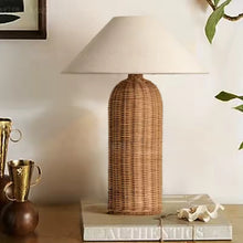 Load image into Gallery viewer, Rattan table lamp on a wooden surface with decorative items. The Yumi Japanese Table Lamp by Allthingscurated embraces the wabi-sabi philosophy that profound beauty emerges from simplicity. Crafted from natural rattan, its minimalist design feels both rustic and contemporary. It elevates your home décor by transforming any space into a sanctuary of quiet sophistication.