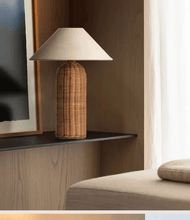 Load image into Gallery viewer, Table lamp with rattan base and beige shade on a wooden shelf. The Yumi Japanese Table Lamp by Allthingscurated embraces the wabi-sabi philosophy that profound beauty emerges from simplicity. Crafted from natural rattan, its minimalist design feels both rustic and contemporary. It elevates your home décor by transforming any space into a sanctuary of quiet sophistication.