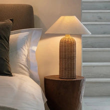 Load image into Gallery viewer, Rattan table lamp on a wooden stump next to a bed with pillows. The Yumi Japanese Table Lamp by Allthingscurated embraces the wabi-sabi philosophy that profound beauty emerges from simplicity. Crafted from natural rattan, its minimalist design feels both rustic and contemporary. It elevates your home décor by transforming any space into a sanctuary of quiet sophistication.