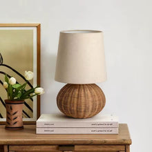 Load image into Gallery viewer, Rattan table lamp with beige shade on a wooden surface with books and a vase of flowers. The Yumi Japanese Table Lamp by Allthingscurated embraces the wabi-sabi philosophy that profound beauty emerges from simplicity. Crafted from natural rattan, its minimalist design feels both rustic and contemporary. It elevates your home décor by transforming any space into a sanctuary of quiet sophistication.