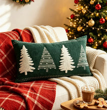 Load image into Gallery viewer, A rectangular green Christmas decor pillow embroidered with four white trees, resting on a sofa with a red plaid blanket and a mug of hot cocoa nearby.
