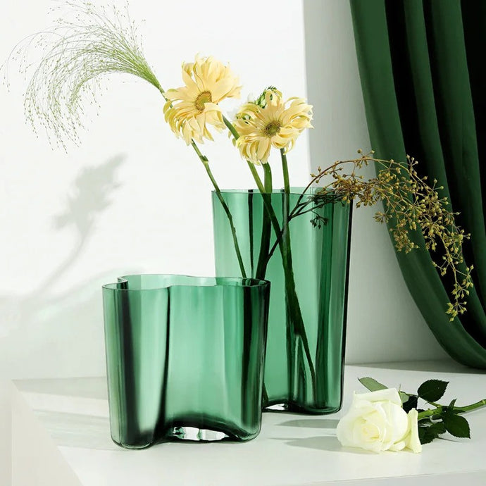 Two green glass vases with flowers on a white surface. The Dayton Vase by Allthingscurated offers a beautiful asymmetrical design that feels both timeless and organic. Its sculptural, minimalist form brings a striking yet harmonious touch to any home interior. Available in clear, green and amber, it’s an unforgettable gift for housewarmings, weddings or holidays.