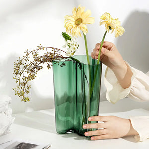 Person arranging flowers into a green vase on a white surface. The Dayton Vase by Allthingscurated offers a beautiful asymmetrical design that feels both timeless and organic. Its sculptural, minimalist form brings a striking yet harmonious touch to any home interior. Available in clear, green and amber, it’s an unforgettable gift for housewarmings, weddings or holidays.