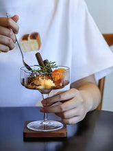 Load image into Gallery viewer, Person holding a glass dish with a dessert, garnished with cinnamon sticks and rosemary. Florentina Drinkware by Allthingscurated embraces a modern design featuring sleek fluted lines with a unique shape reminiscent of a peach blossom flower. Stylish and chic, this collection includes a carafe, goblets and several tumblers, including a glamorous coupe cocktail glass. Add elegance to everyday use with this beautiful collection and just as perfect for setting an extravagant holiday table.
