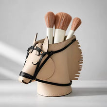 Load image into Gallery viewer, A horse-shaped makeup brush holder in beige faux leather with black bridle details, holding several makeup brushes, styled on a textured book against a neutral background.
