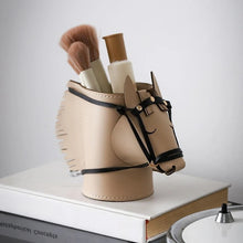 Load image into Gallery viewer, A beige horse-shaped makeup brush holder with black leather-like straps and trim, filled with makeup brushes and a cosmetic tube, resting on a white book against a light gray background.
