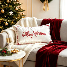 Load image into Gallery viewer, A rectangular white pillow with red Merry Christmas embroidery rests on a couch with a red throw blanket, creating a festive mood next to a small table with a glass of champagne and gold chocolates.
