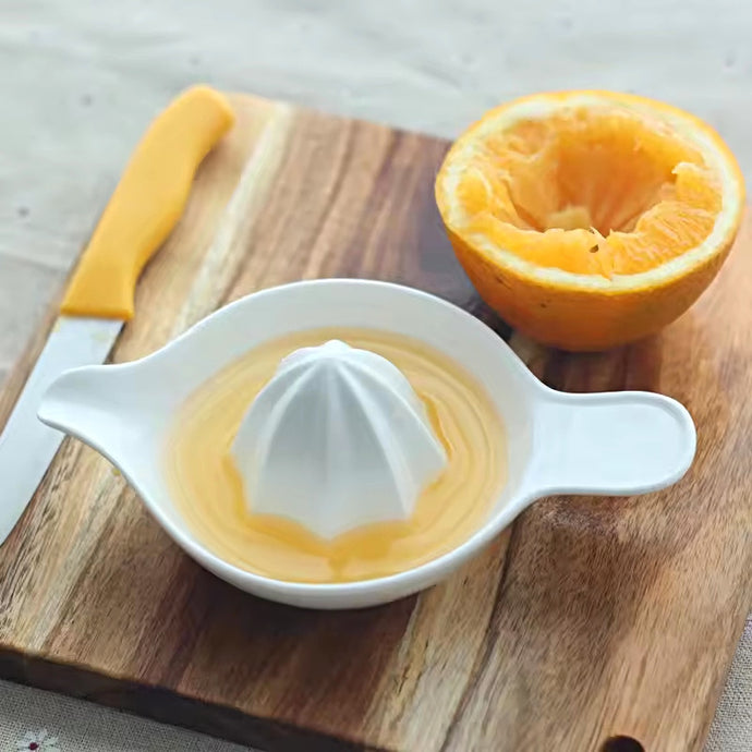 White manual juicer with freshly squeezed orange juice on a wooden cutting board. Ceramic Citrus Juicer by Allthingscurated features a classic reamer design with built-in pour spout that ensures precise pouring without spills. This single-piece tool is easy to store and indispensable in any bar or kitchen.