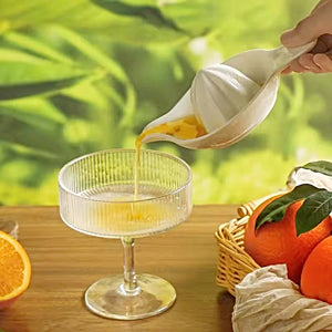 Person pouring orange juice into a cocktail glass with oranges and a basket in the background. Ceramic Citrus Juicer by Allthingscurated features a classic reamer design with built-in pour spout that ensures precise pouring without spills. This single-piece tool is easy to store and indispensable in any bar or kitchen.