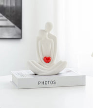 Load image into Gallery viewer, This romantic sculpture by Allthingscurated elegantly portrays a seated couple, locked in a tender embrace, their love and hearts held close. Sculpted in white ceramic, the piece highlights a striking red heart that symbolizes deep connection and passion. An ideal gift for Valentine’s Day or weddings, it offers a refined and timeless expression of affection.

