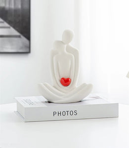 This romantic sculpture by Allthingscurated elegantly portrays a seated couple, locked in a tender embrace, their love and hearts held close. Sculpted in white ceramic, the piece highlights a striking red heart that symbolizes deep connection and passion. An ideal gift for Valentine’s Day or weddings, it offers a refined and timeless expression of affection.