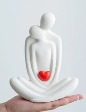 Load image into Gallery viewer, This romantic sculpture by Allthingscurated elegantly portrays a seated couple, locked in a tender embrace, their love and hearts held close. Sculpted in white ceramic, the piece highlights a striking red heart that symbolizes deep connection and passion. An ideal gift for Valentine’s Day or weddings, it offers a refined and timeless expression of affection.
