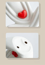 Load image into Gallery viewer, This romantic sculpture by Allthingscurated elegantly portrays a seated couple, locked in a tender embrace, their love and hearts held close. Sculpted in white ceramic, the piece highlights a striking red heart that symbolizes deep connection and passion. An ideal gift for Valentine’s Day or weddings, it offers a refined and timeless expression of affection.
