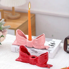 Load image into Gallery viewer, Pink and red bow-shaped candle holders with a lit orange candle on a white surface. Fashion Bow Candle Holder by Allthingscurated spots a distinctive knot design, beautiful yet practical. Perfect for wedding celebrations, home styling, or to infuse warmth and romance into any living space. Available in three colors and 2 sizes.
