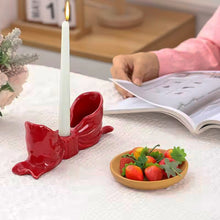 Load image into Gallery viewer, Red bow-shaped candle holder with a lit candle on a table next to a small plate of fruit. Fashion Bow Candle Holder by Allthingscurated spots a distinctive knot design, beautiful yet practical. Perfect for wedding celebrations, home styling, or to infuse warmth and romance into any living space. Available in three colors and 2 sizes. 
