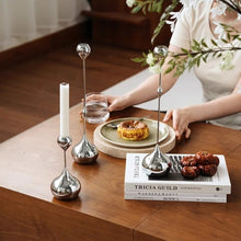 Load image into Gallery viewer, Decorative silver candle holders on a wooden table with books and food. Lumea Silver Candle Holders by Allthingscurated feature a striking trio of sculptural pieces that blend contemporary design with timeless elegance. Each holder features a distinctive water droplet base that anchors a sleek, minimalist silhouette, creating an art deco-inspired statement on its own. this set makes an exceptional wedding gift for couples seeking sophisticated home décor that transcends occasion.
