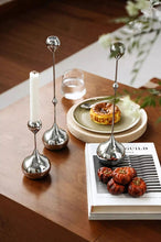 Load image into Gallery viewer, Decorative silver candle holders on a wooden table with a book and small plate. Lumea Silver Candle Holders by Allthingscurated feature a striking trio of sculptural pieces that blend contemporary design with timeless elegance. Each holder features a distinctive water droplet base that anchors a sleek, minimalist silhouette, creating an art deco-inspired statement on its own. this set makes an exceptional wedding gift for couples seeking sophisticated home décor that transcends occasion.
