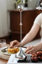 Load image into Gallery viewer, Silver candle holder on a table with a blurred background. Lumea Silver Candle Holders by Allthingscurated feature a striking trio of sculptural pieces that blend contemporary design with timeless elegance. Each holder features a distinctive water droplet base that anchors a sleek, minimalist silhouette, creating an art deco-inspired statement on its own. this set makes an exceptional wedding gift for couples seeking sophisticated home décor that transcends occasion.
