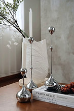 Load image into Gallery viewer, Decorative silver candle holders on a table with books and a candle. Lumea Silver Candle Holders by Allthingscurated feature a striking trio of sculptural pieces that blend contemporary design with timeless elegance. Each holder features a distinctive water droplet base that anchors a sleek, minimalist silhouette, creating an art deco-inspired statement on its own. this set makes an exceptional wedding gift for couples seeking sophisticated home décor that transcends occasion.
