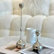 Load image into Gallery viewer, Two silver candle holders on a table with a blurred background. Lumea Silver Candle Holders by Allthingscurated feature a striking trio of sculptural pieces that blend contemporary design with timeless elegance. Each holder features a distinctive water droplet base that anchors a sleek, minimalist silhouette, creating an art deco-inspired statement on its own. this set makes an exceptional wedding gift for couples seeking sophisticated home décor that transcends occasion.
