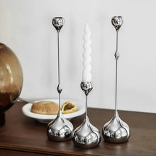 Load image into Gallery viewer, Three silver candle holders with a white candle on a wooden surface. Lumea Silver Candle Holders by Allthingscurated feature a striking trio of sculptural pieces that blend contemporary design with timeless elegance. Each holder features a distinctive water droplet base that anchors a sleek, minimalist silhouette, creating an art deco-inspired statement on its own. this set makes an exceptional wedding gift for couples seeking sophisticated home décor that transcends occasion.
