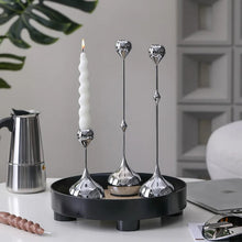 Load image into Gallery viewer, Decorative candle holders on a black tray with a lit candle, placed on a desk with office items. Lumea Silver Candle Holders by Allthingscurated feature a striking trio of sculptural pieces that blend contemporary design with timeless elegance. Each holder features a distinctive water droplet base that anchors a sleek, minimalist silhouette, creating an art deco-inspired statement on its own. this set makes an exceptional wedding gift for couples seeking sophisticated home décor that transcends occasion.
