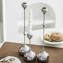 Load image into Gallery viewer, Three silver candle holders with a white candle on a table. Lumea Silver Candle Holders by Allthingscurated feature a striking trio of sculptural pieces that blend contemporary design with timeless elegance. Each holder features a distinctive water droplet base that anchors a sleek, minimalist silhouette, creating an art deco-inspired statement on its own. this set makes an exceptional wedding gift for couples seeking sophisticated home décor that transcends occasion.
