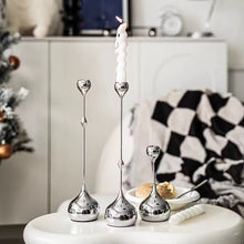 Load image into Gallery viewer, Silver candle holders with candles on a table in a home setting. Lumea Silver Candle Holders by Allthingscurated feature a striking trio of sculptural pieces that blend contemporary design with timeless elegance. Each holder features a distinctive water droplet base that anchors a sleek, minimalist silhouette, creating an art deco-inspired statement on its own. this set makes an exceptional wedding gift for couples seeking sophisticated home décor that transcends occasion.
