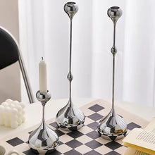 Load image into Gallery viewer, Set of three silver candle holders on a checkered surface with a white curtain background. Lumea Silver Candle Holders by Allthingscurated feature a striking trio of sculptural pieces that blend contemporary design with timeless elegance. Each holder features a distinctive water droplet base that anchors a sleek, minimalist silhouette, creating an art deco-inspired statement on its own. this set makes an exceptional wedding gift for couples seeking sophisticated home décor that transcends occasion.
